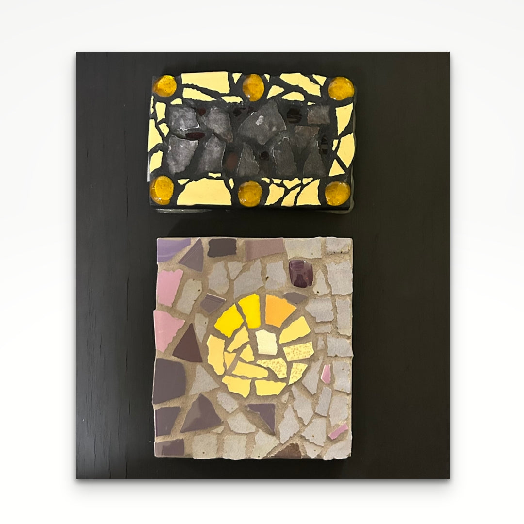 Two panel mosaics of yellow and black tiles, one above the other, on a plank of wood painted black. 