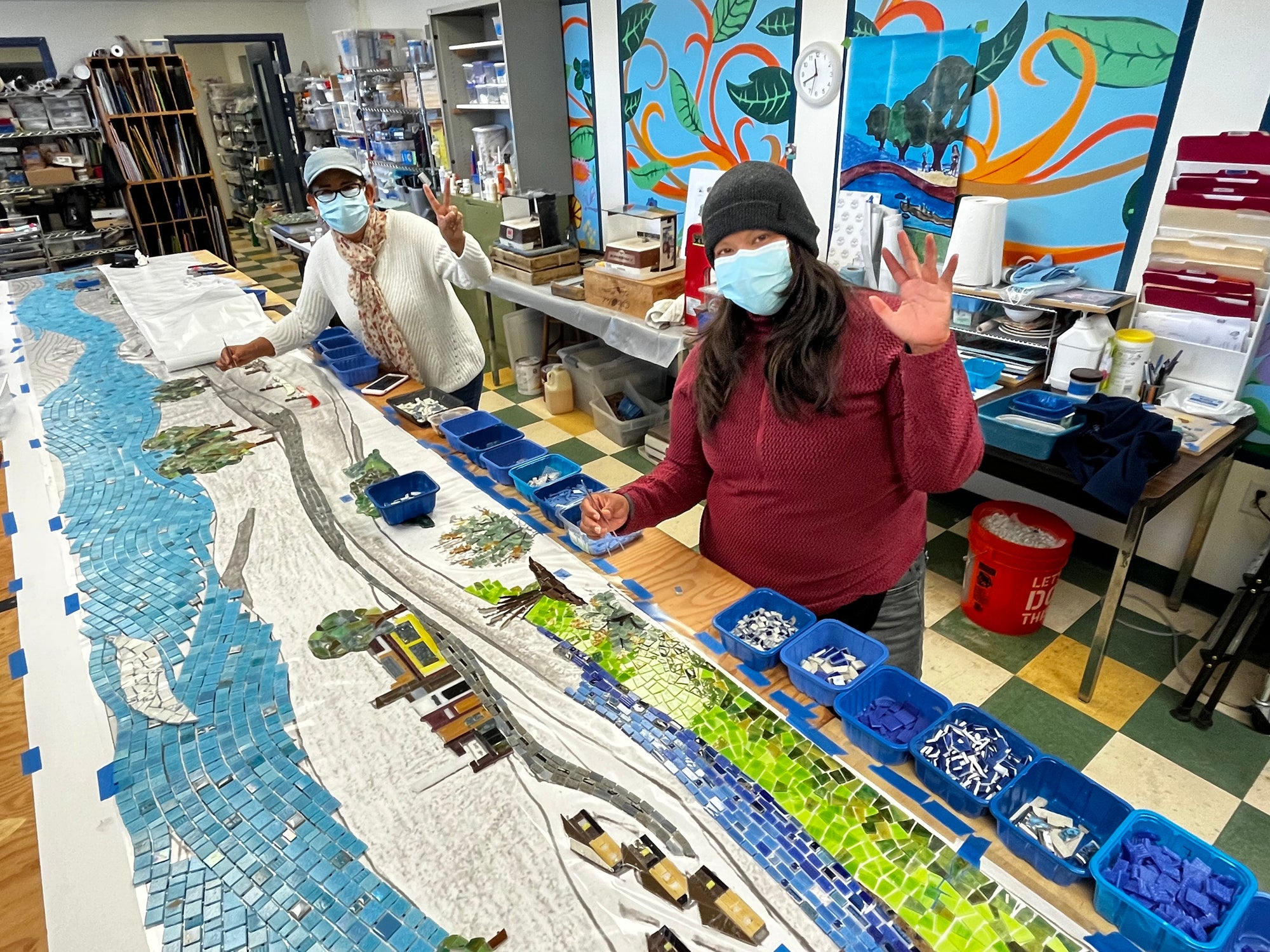 Artists working on a large-scale mosaic and waving to the camera.