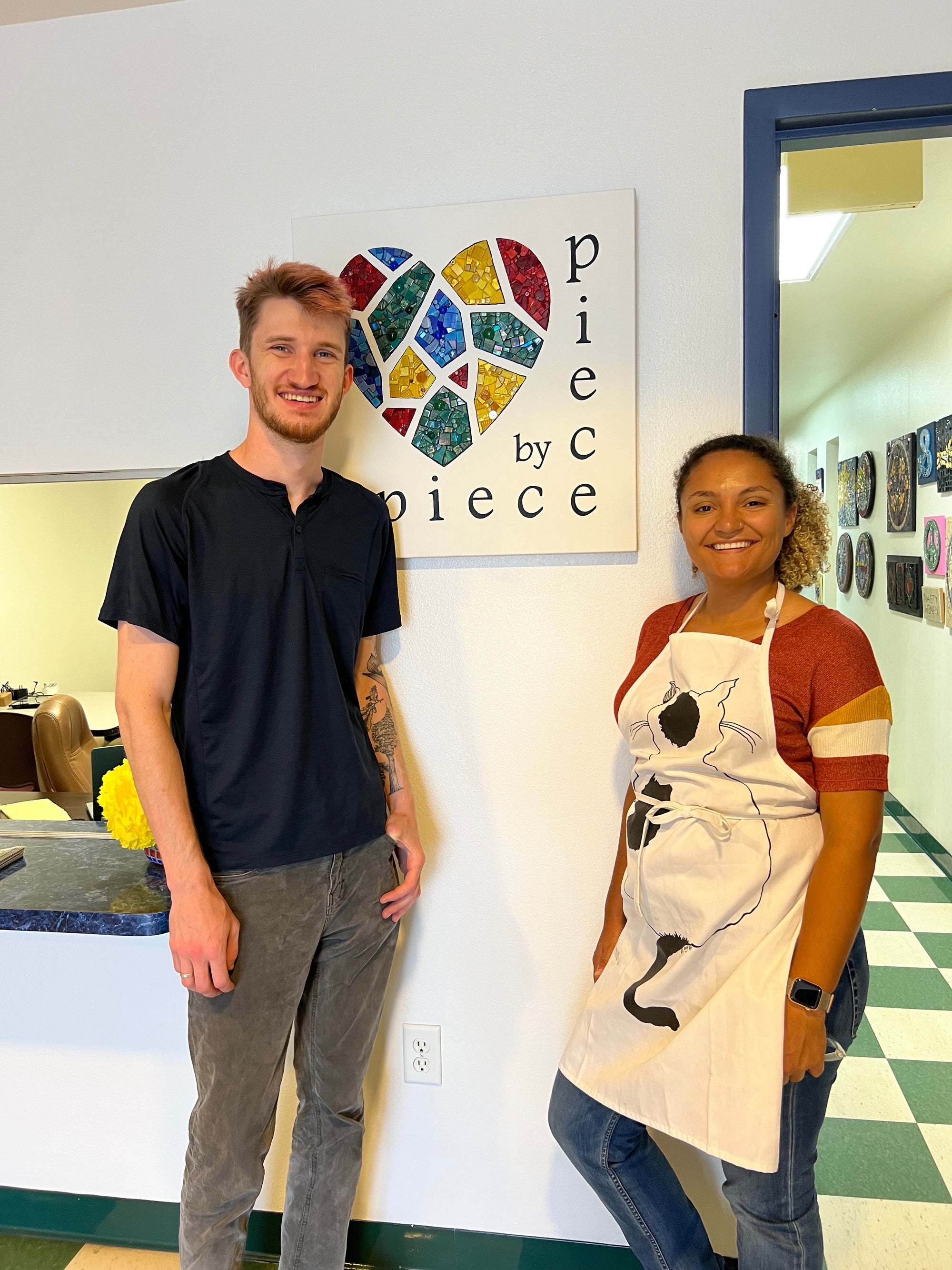 Two new employees stand near a mosaic of the Piece by Piece logo.