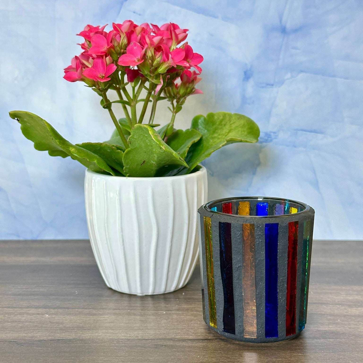 Dark colored vertical line mosaic votive on a table next to a planter with pink flowers.