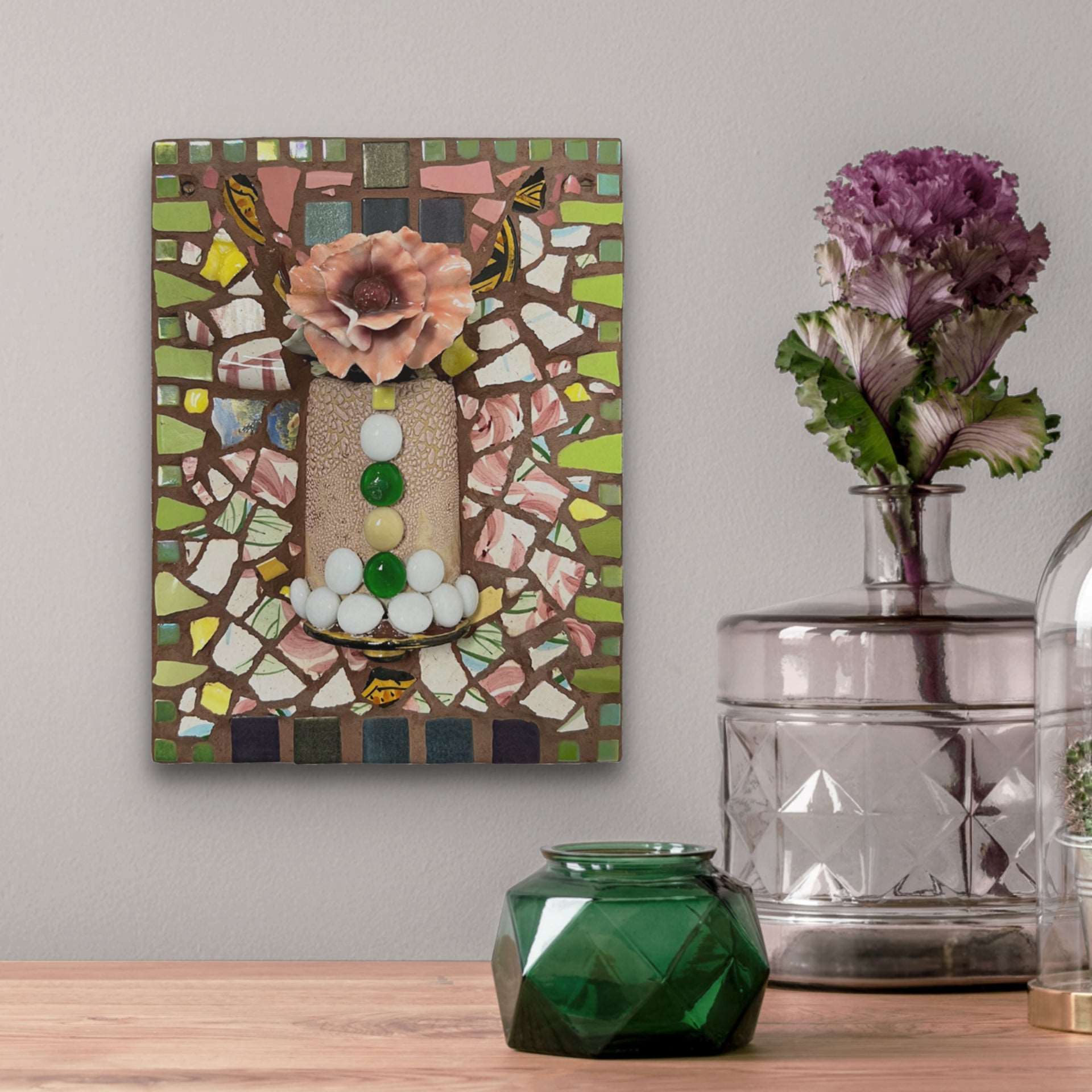 A pink ceramic flower sits atop a vessel that is set in a field of pink and white mosaic pieces with a beautiful green border.